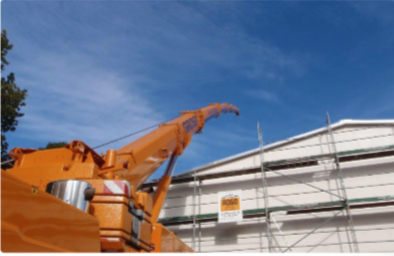 Ein Kran hebt Baumaterial vor einem Gerüst an einem Gebäude unter blauem Himmel.
