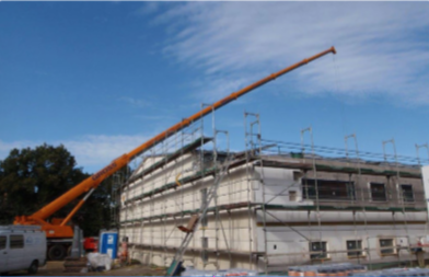 Baustelle mit Gebäudegerüst, Kran im Einsatz. Blau-weißer Himmel im Hintergrund. Haus in Bauphase, Baum links im Bild.