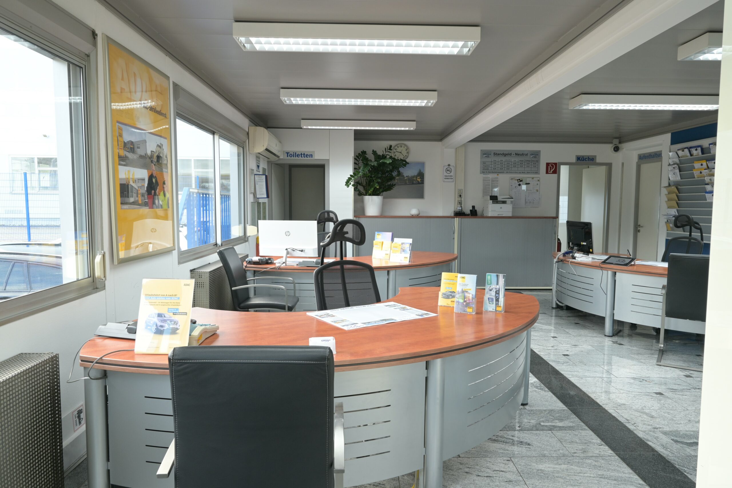 Modernes Büro mit mehreren stilvollen Holztischen, Computern und Broschüren, umgeben von großen Fenstern mit Licht.