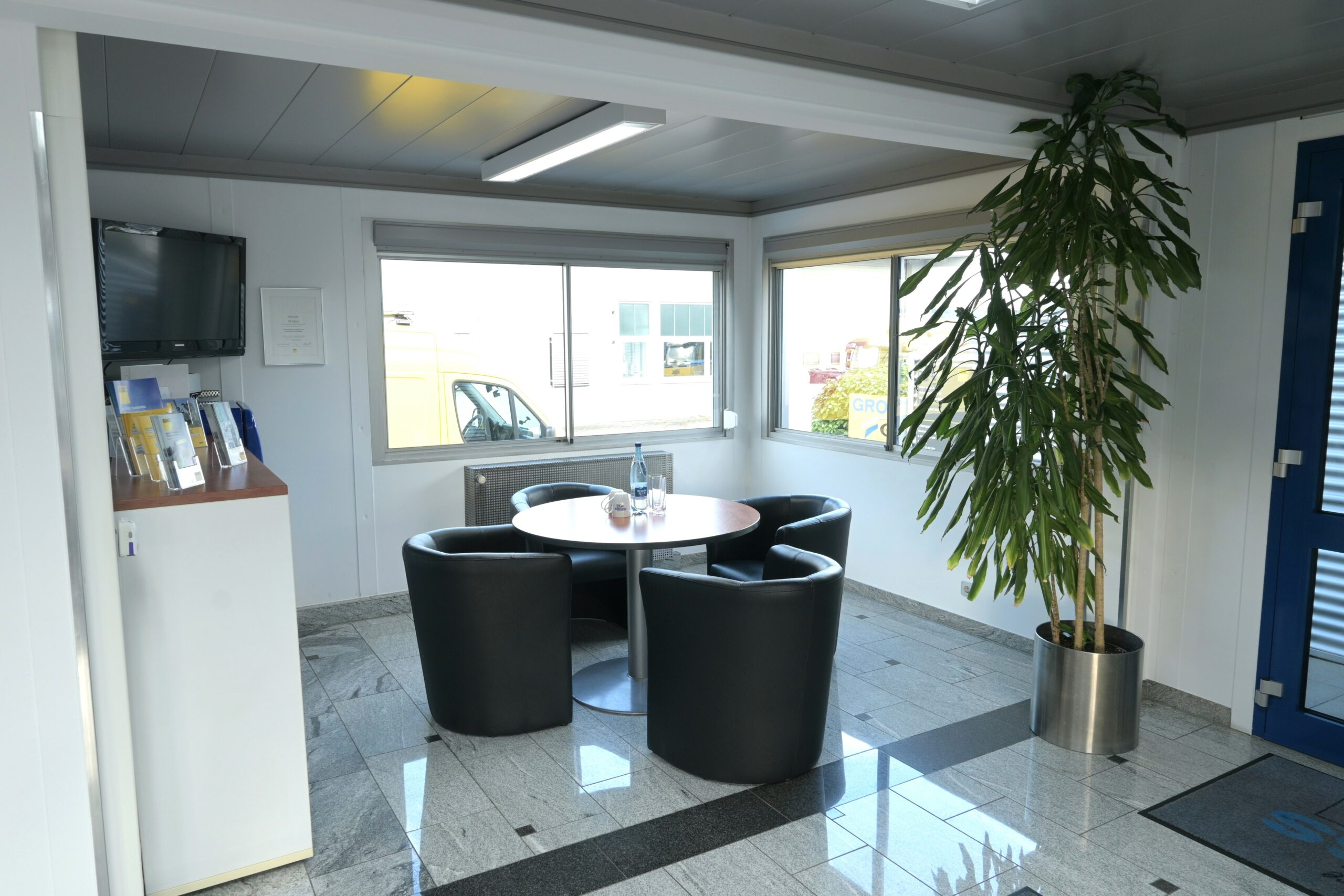Moderner Warteraum mit schwarzen Sesseln um runden Tisch, großen Fenstern, Topfpflanze und Broschürenständer.