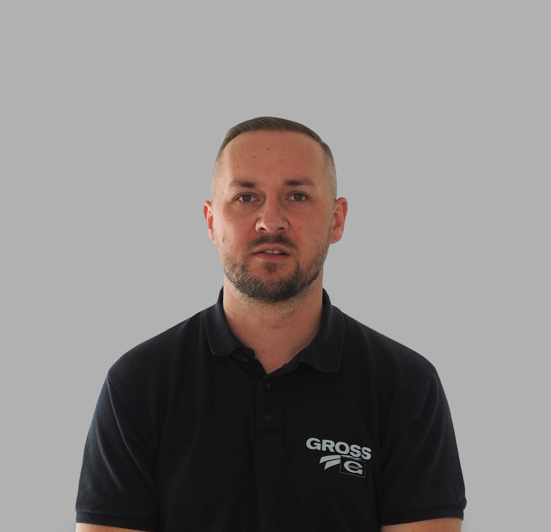 Ein Mann mit Bart trägt ein schwarzes Polohemd mit Logo vor grauem Hintergrund.