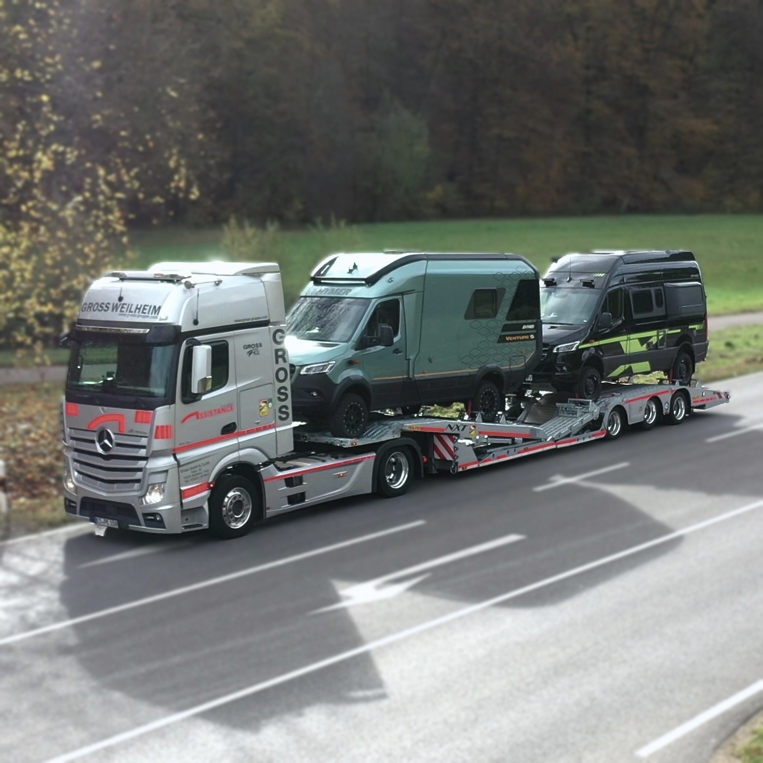 Lkw mit zwei Wohnmobilen auf einem Anhänger fährt auf einer Landstraße mit grüner Landschaft im Hintergrund.