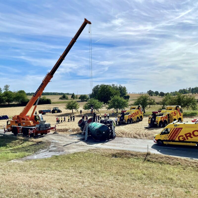 Kran hebt umgekippten Tanklastwagen auf einem Feld, umgeben von Bergungsfahrzeugen und Menschen, bei sonnigem Wetter.