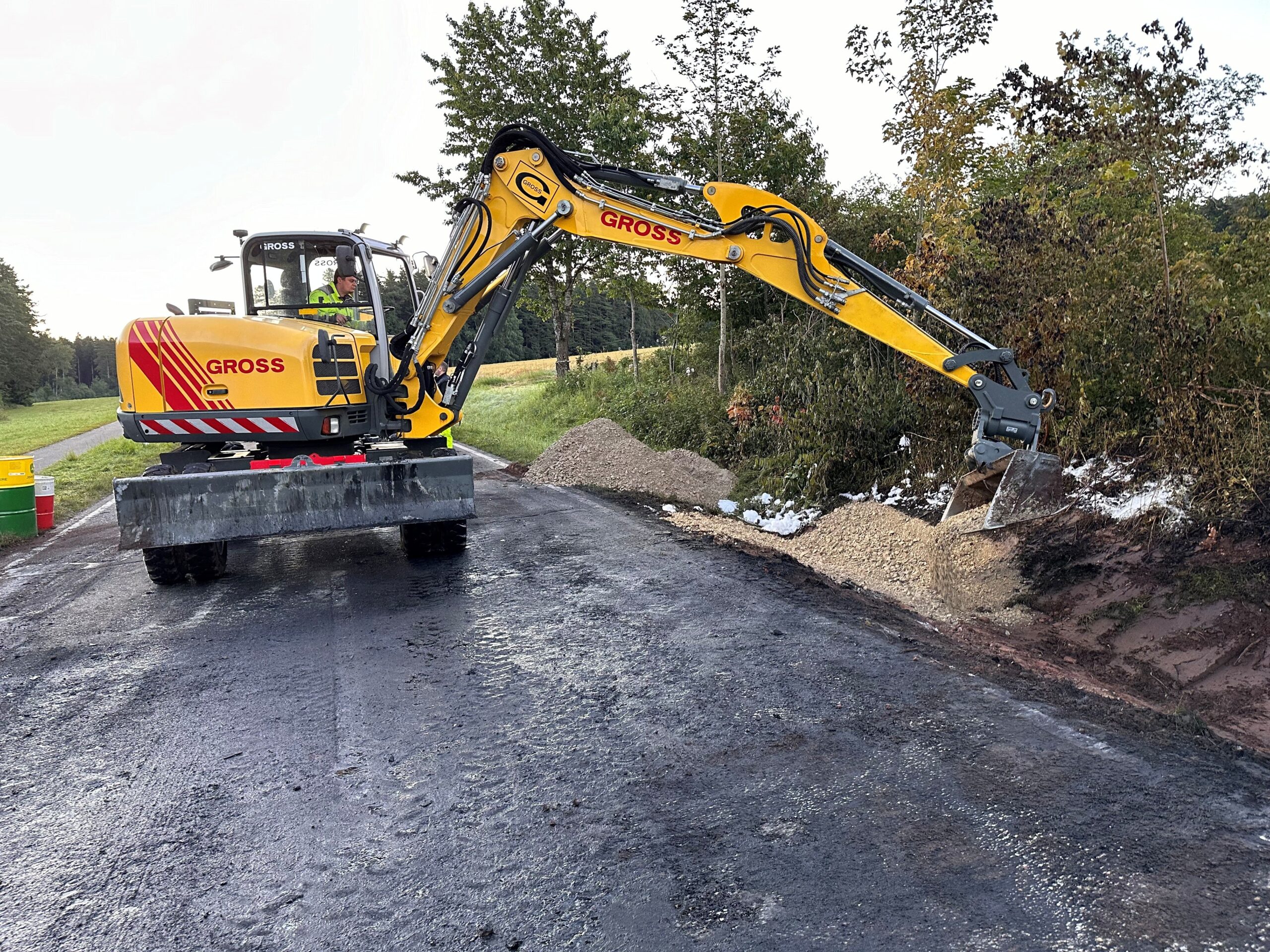 Bagger auf Straße hebt Kieshaufen neben Wald auf. Baustelle auf Asphalt unter freiem Himmel mit hellen und dunklen Bodenflächen.