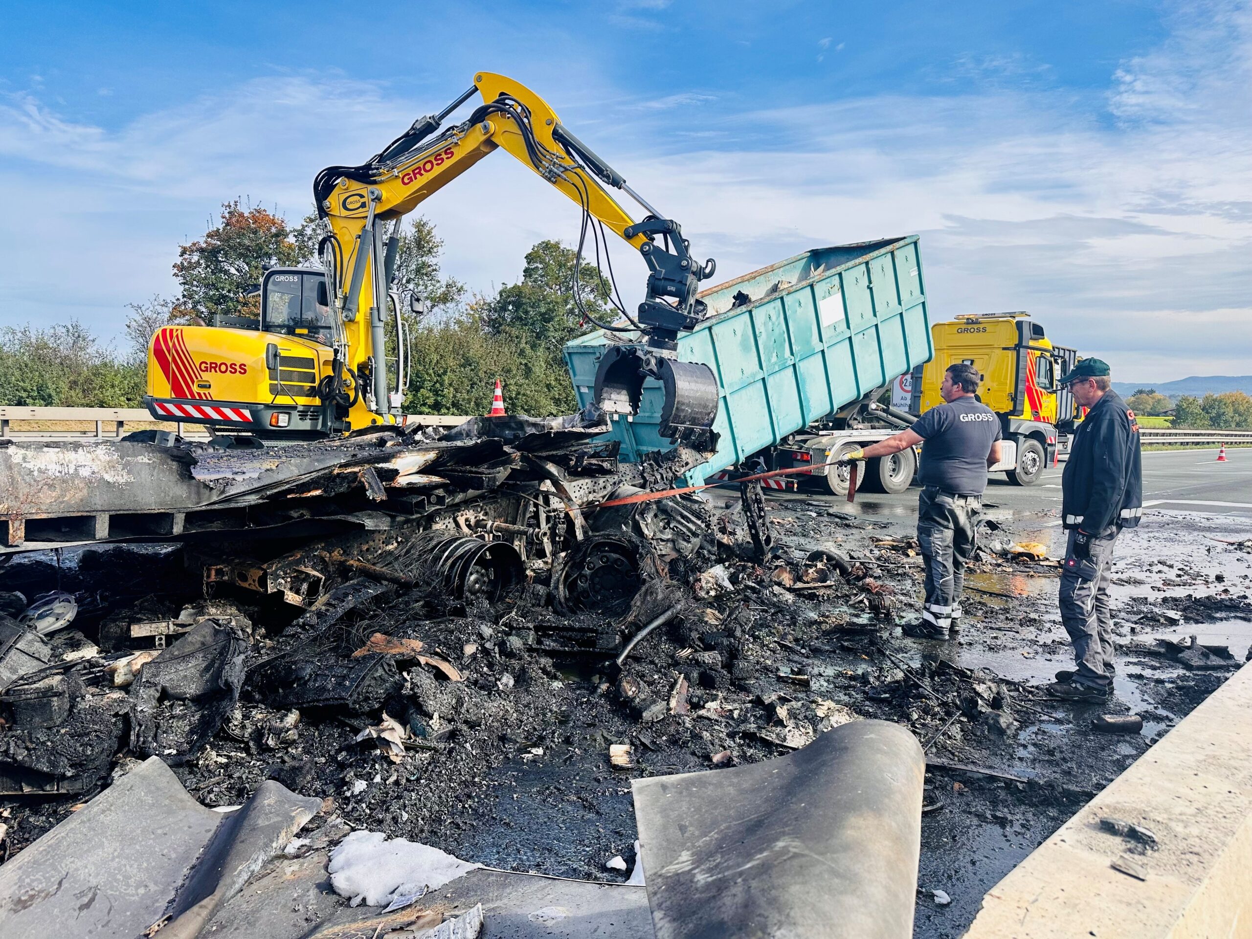 Ein gelber Bagger räumt verbrannte Trümmer auf einer Autobahn, während daneben zwei Männer den Einsatz koordinieren.
