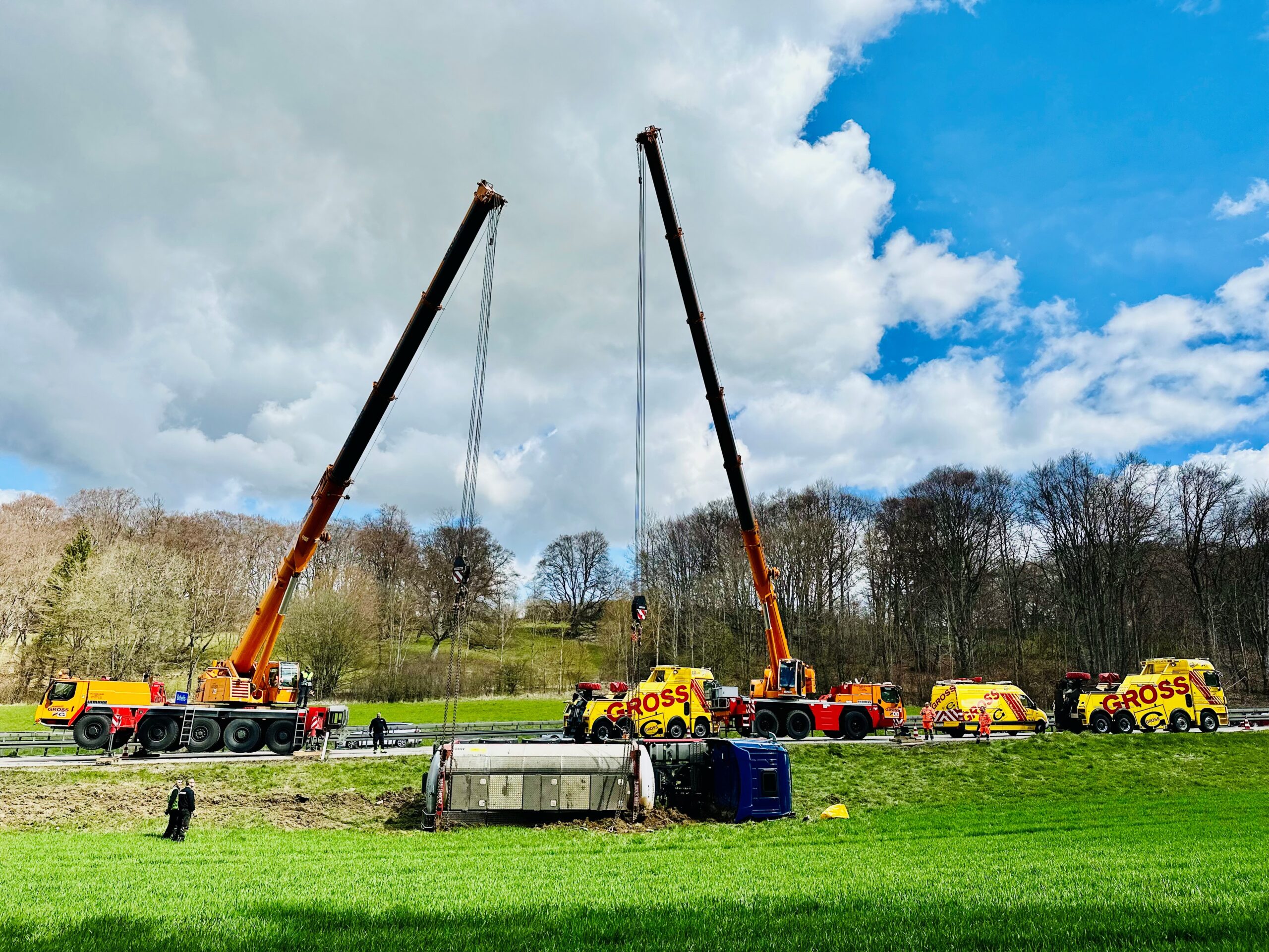 Zwei Kräne heben umgestürzten LKW auf Wiese neben Autobahn, umgeben von Bäumen und bewölktem Himmel.