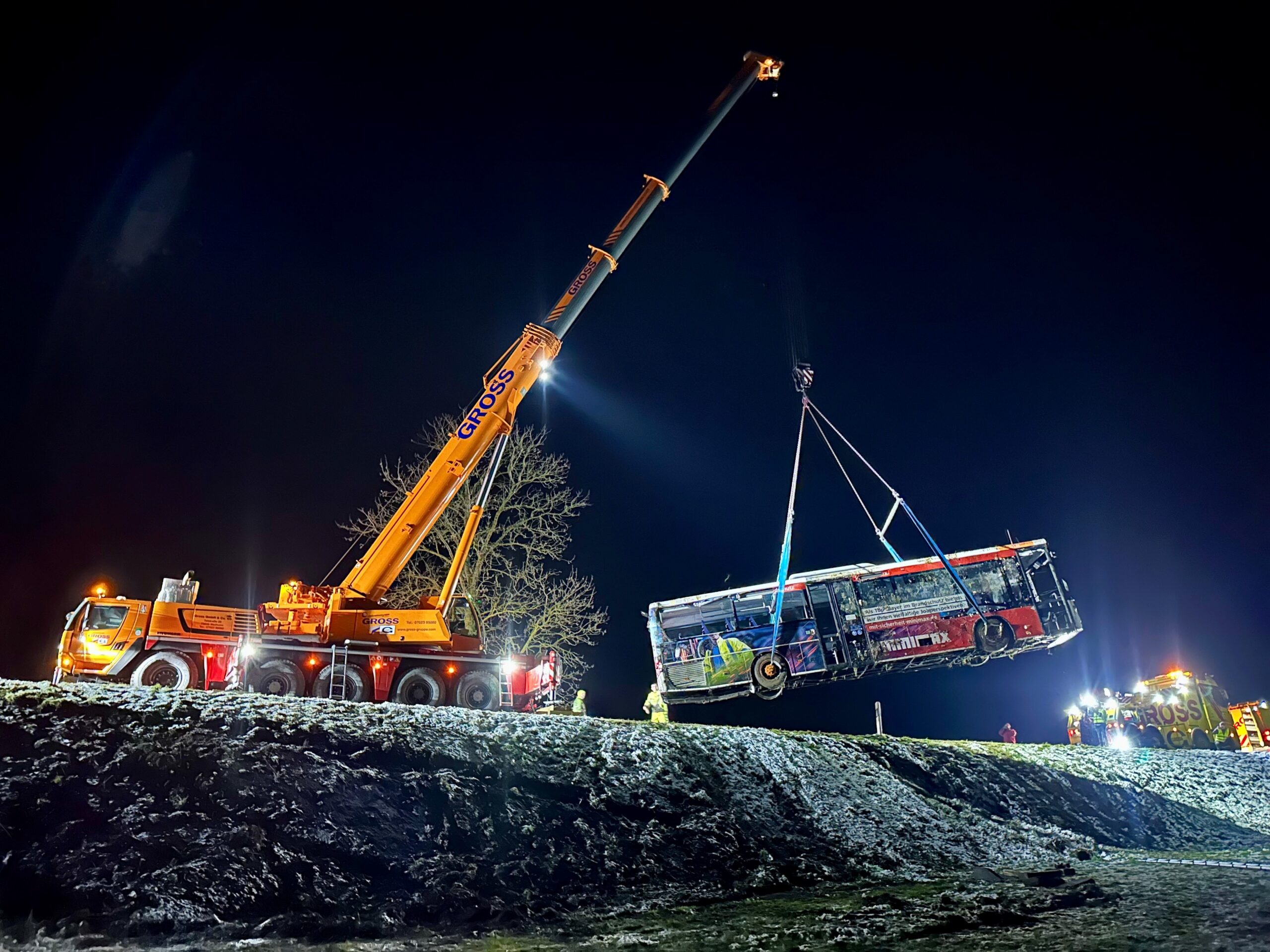 Nächtlicher Einsatz: Ein orangefarbener Kran hebt einen beschädigten Bus aus einem Graben, umgeben von Helfern und Scheinwerfern.