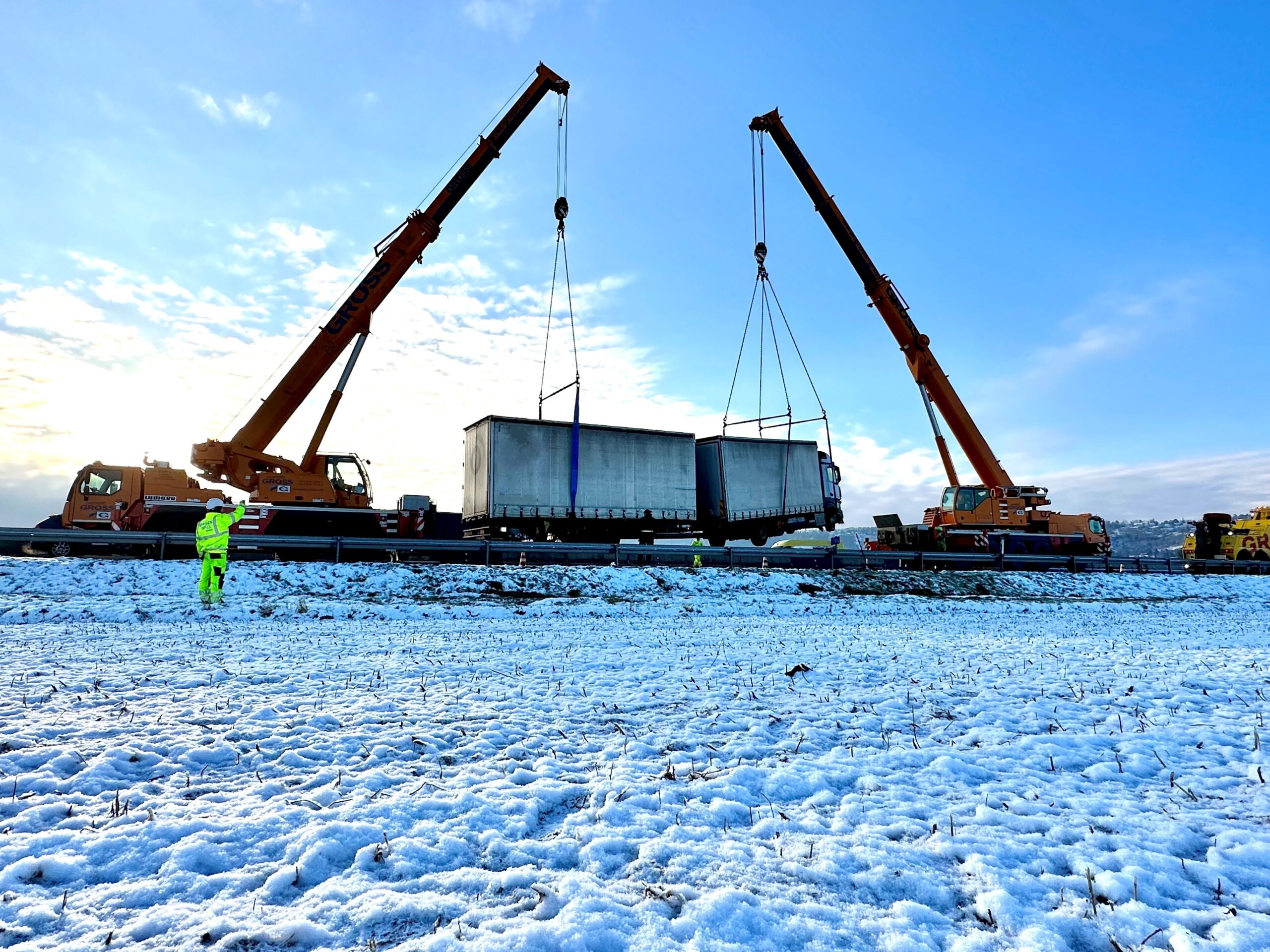 Zwei Kräne heben bei Schneebedeckung einen Lkw-Anhänger an. Ein Arbeiter in leuchtender Kleidung überwacht die Szene.