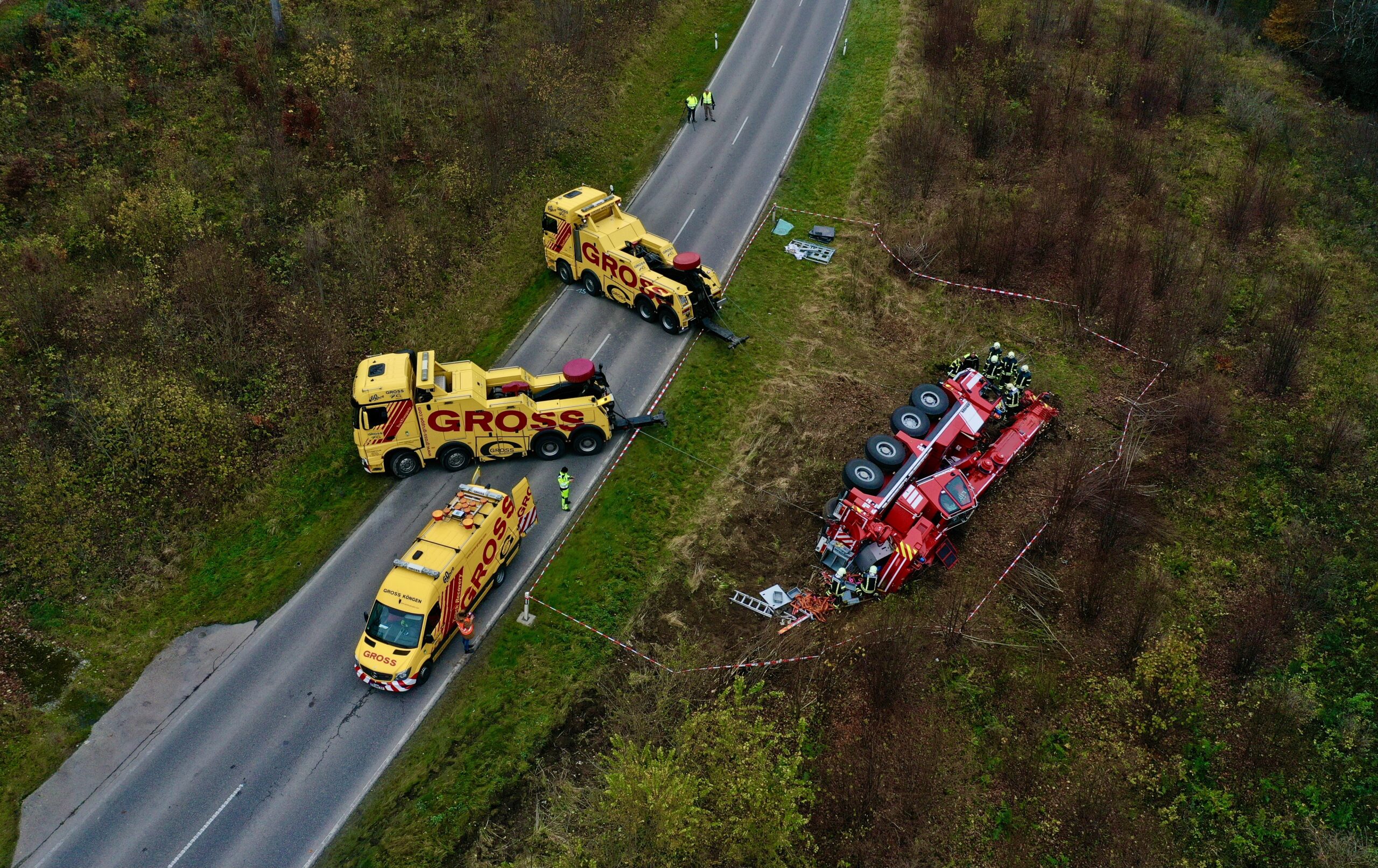 Zwei gelbe Abschleppwagen stehen auf der Straße neben einem umgestürzten roten Lastwagen im Graben. Arbeiter sichern den Bereich.