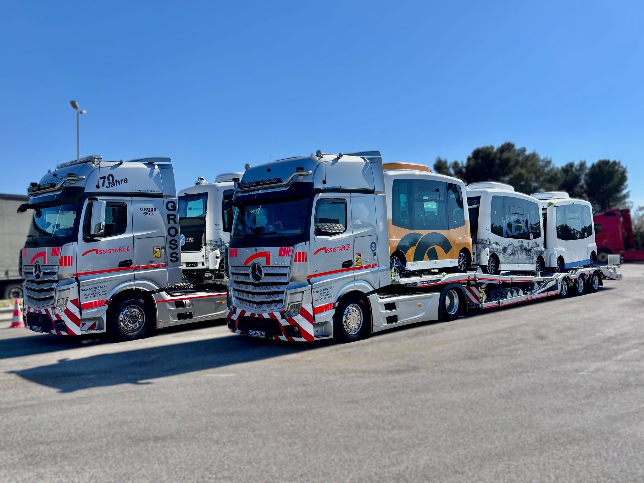 Zwei Lastwagen transportieren autonome Kleinbusse auf einem sonnigen Parkplatz vor Bäumen, Himmel ist klar und blau.