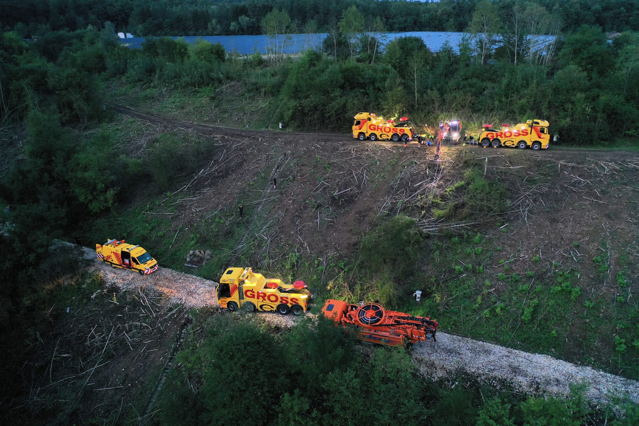 Gelbe Abschleppwagen ziehen ein großes Fahrzeug durch einen bewaldeten Abhang; Abenddämmerung und ruhige Landschaft.