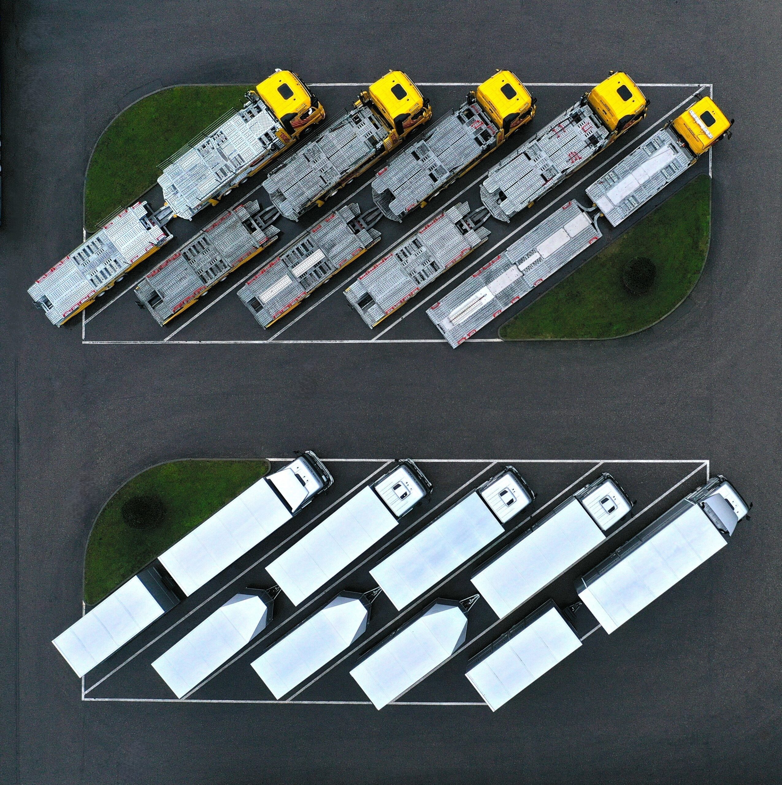 Luftaufnahme: Mehrere gelbe Lastwagen und weiße Anhänger sind geordnet in zwei Reihen auf einem asphaltierten Parkplatz abgestellt.