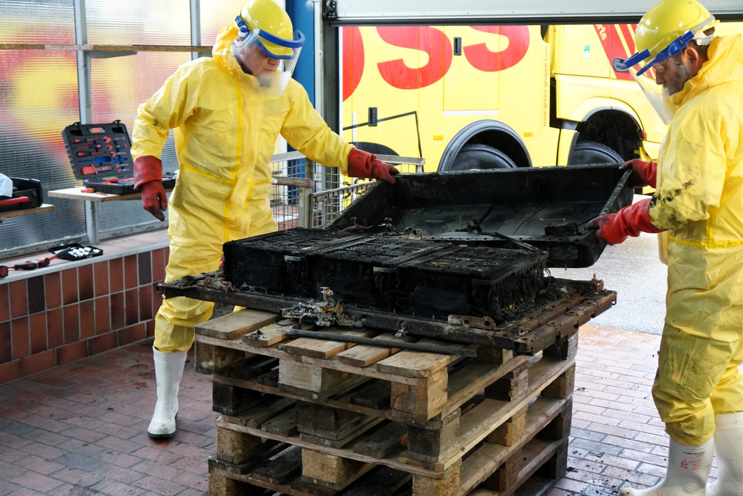 Zwei Personen in Schutzanzügen demontieren eine verbrannte Batterie auf Paletten in einer Werkstatt.