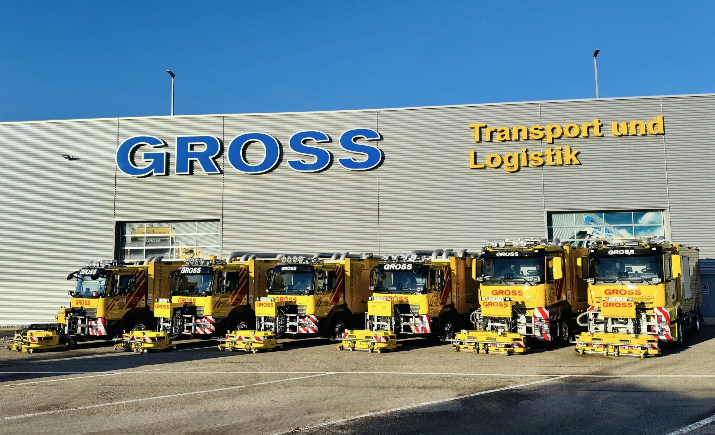 Sechs gelbe Groß-Transportfahrzeuge parken vor einem grauen Gebäude mit dem Schriftzug Gross Transport und Logistik.