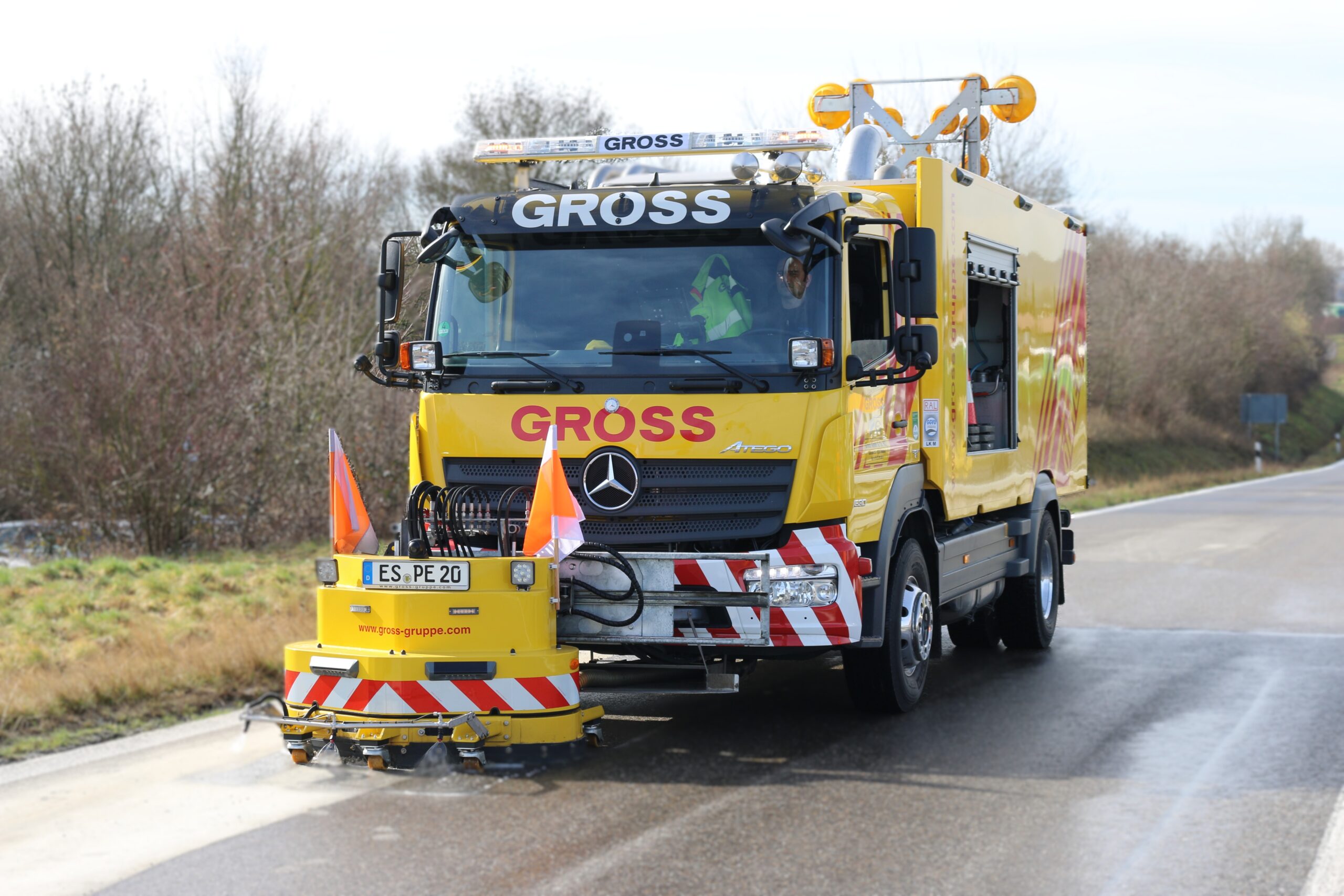 Gelbes Straßenreinigungsfahrzeug mit GROß-Logo sprüht Wasser auf nasser Straße, begleitet von orangen Sicherheitsfahnen.