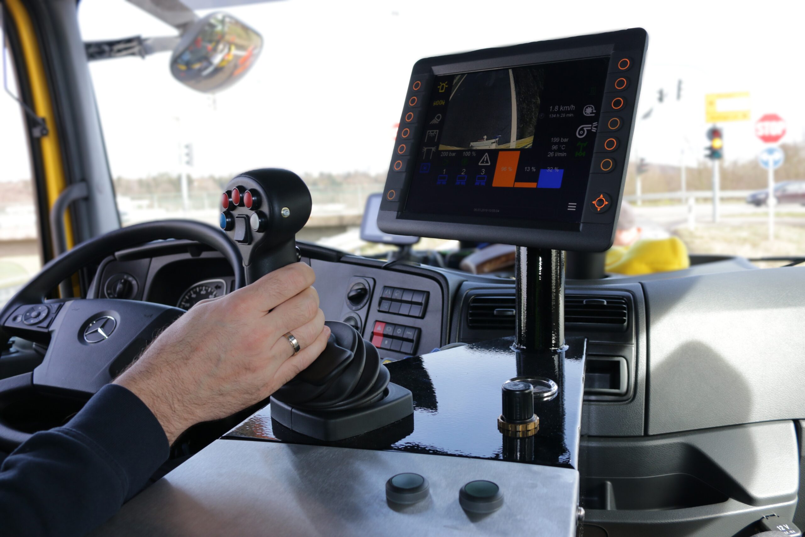 Mann steuert Lkw mit Joystick, während ein Bildschirm Fahrdaten anzeigt. Lenkrad und Außenspiegel sind sichtbar.