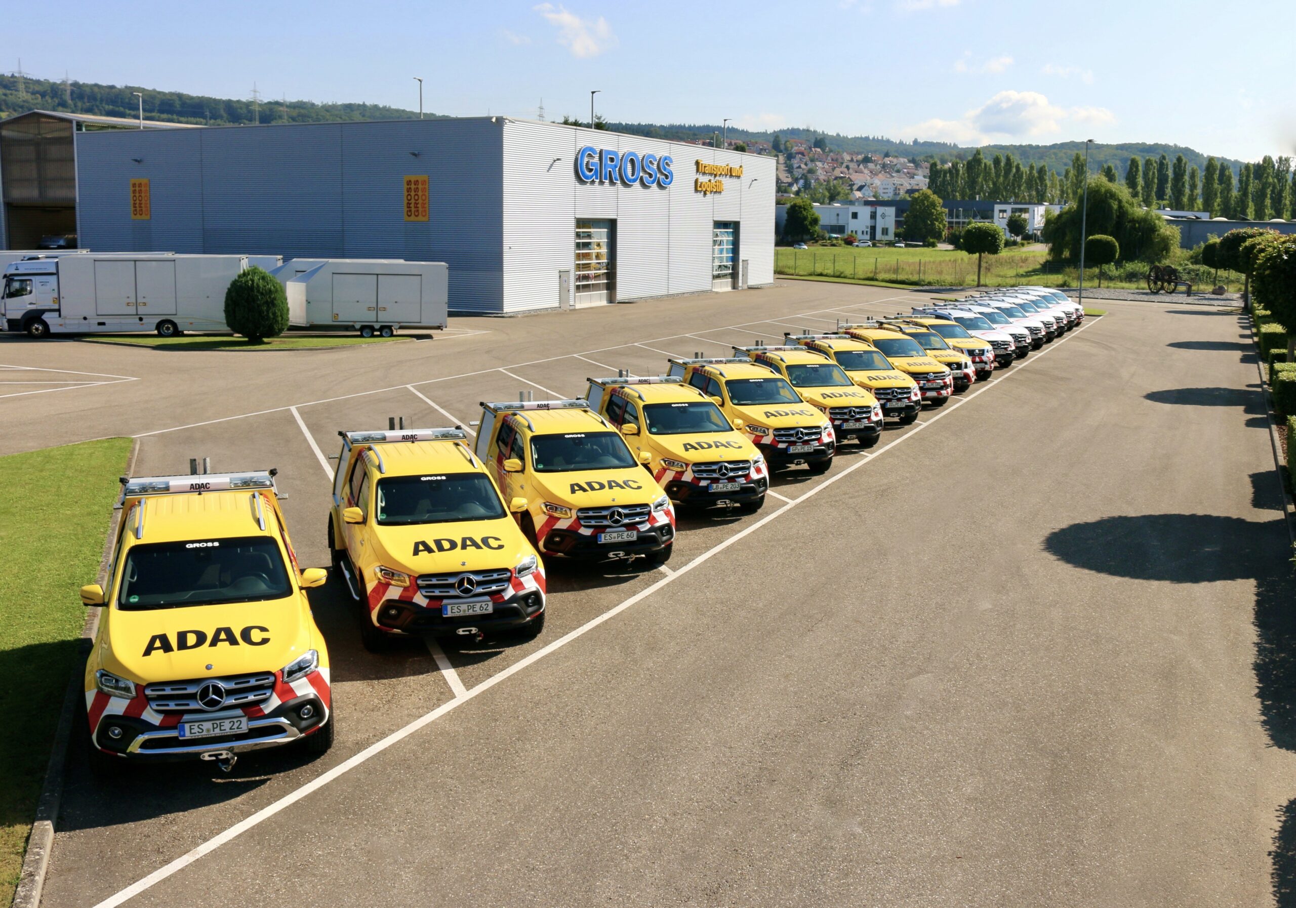 Reihe gelber ADAC-Fahrzeuge parkt vor Groß-Transport-Logistikgebäude, sonniger Tag, leicht hügelige Landschaft im Hintergrund.