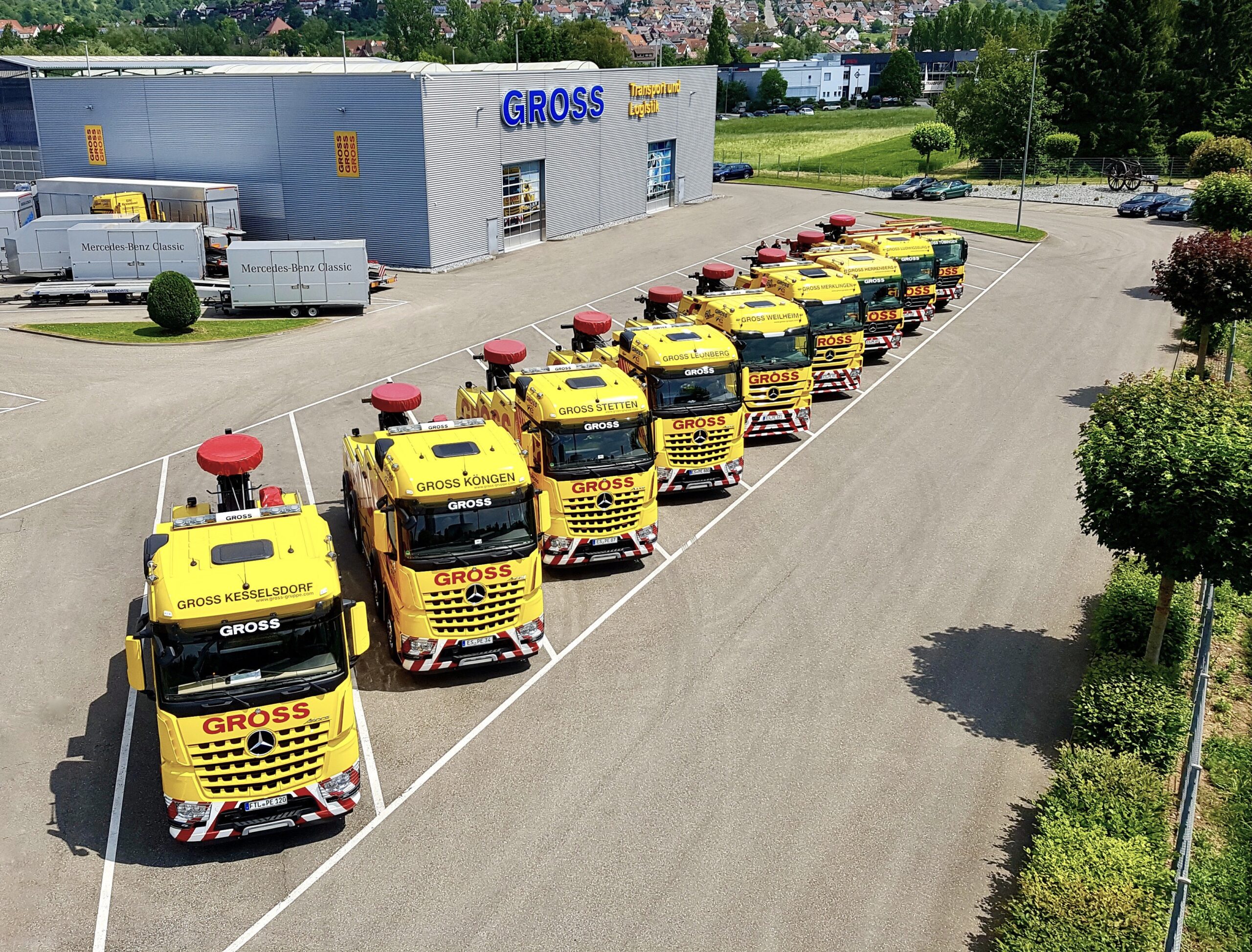 Gelbe Lastwagen von GROSS stehen in einer Reihe vor einem großen Gebäude mit dem Schriftzug „GROSS“, umgeben von Grünflächen.