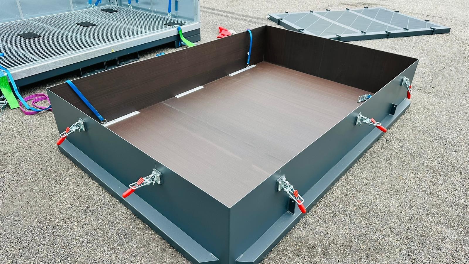 Großer, rechteckiger Metallrahmen mit Hebelverschlüssen auf Asphalt; seitlich bunte Gurte und Gitterflächen.