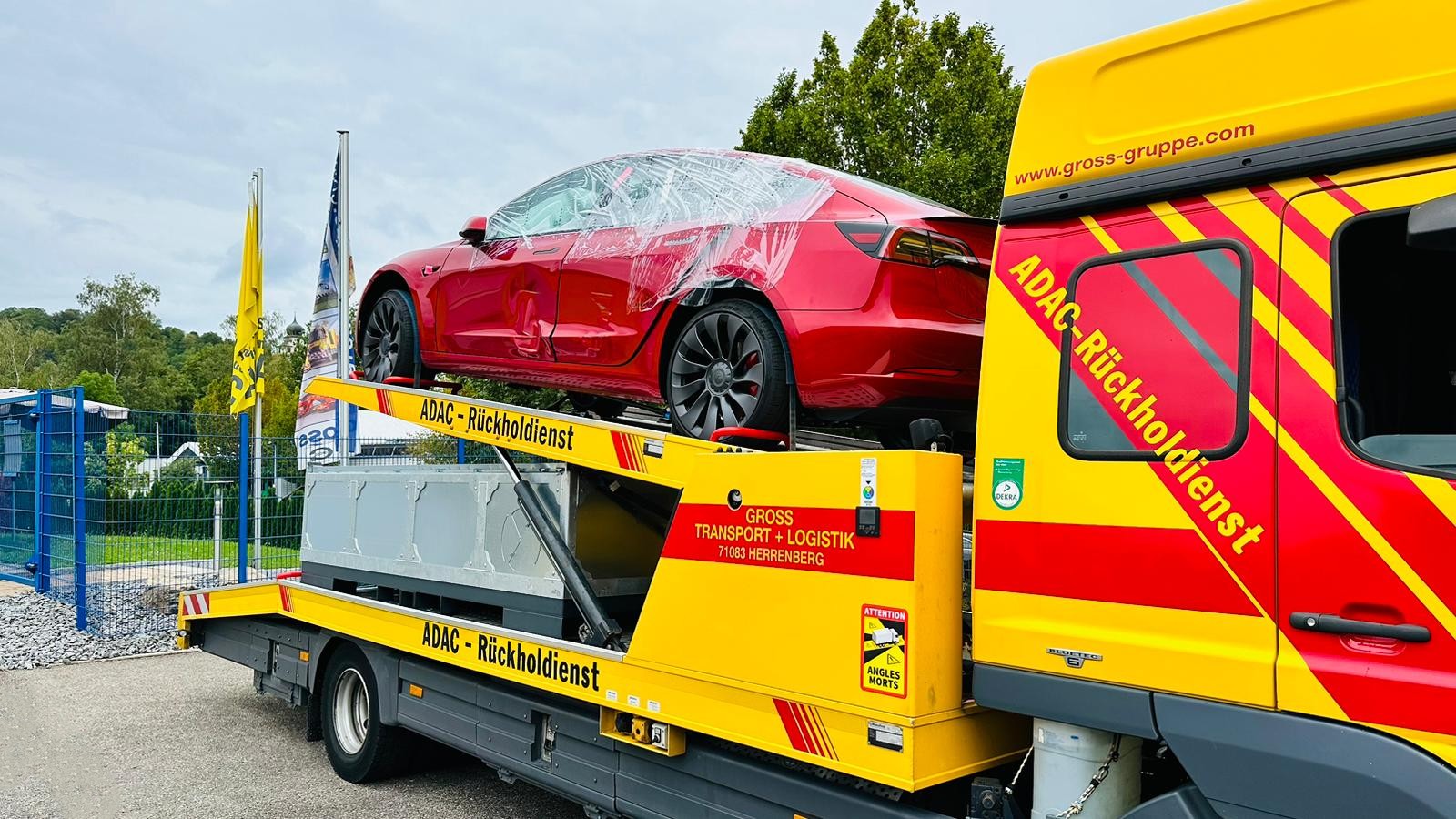 Rotes Auto auf gelbem ADAC-Abschleppfahrzeug, Seitenansicht. Auto teils mit Schutzfolie bedeckt. Hintergründe mit blauem Zaun.