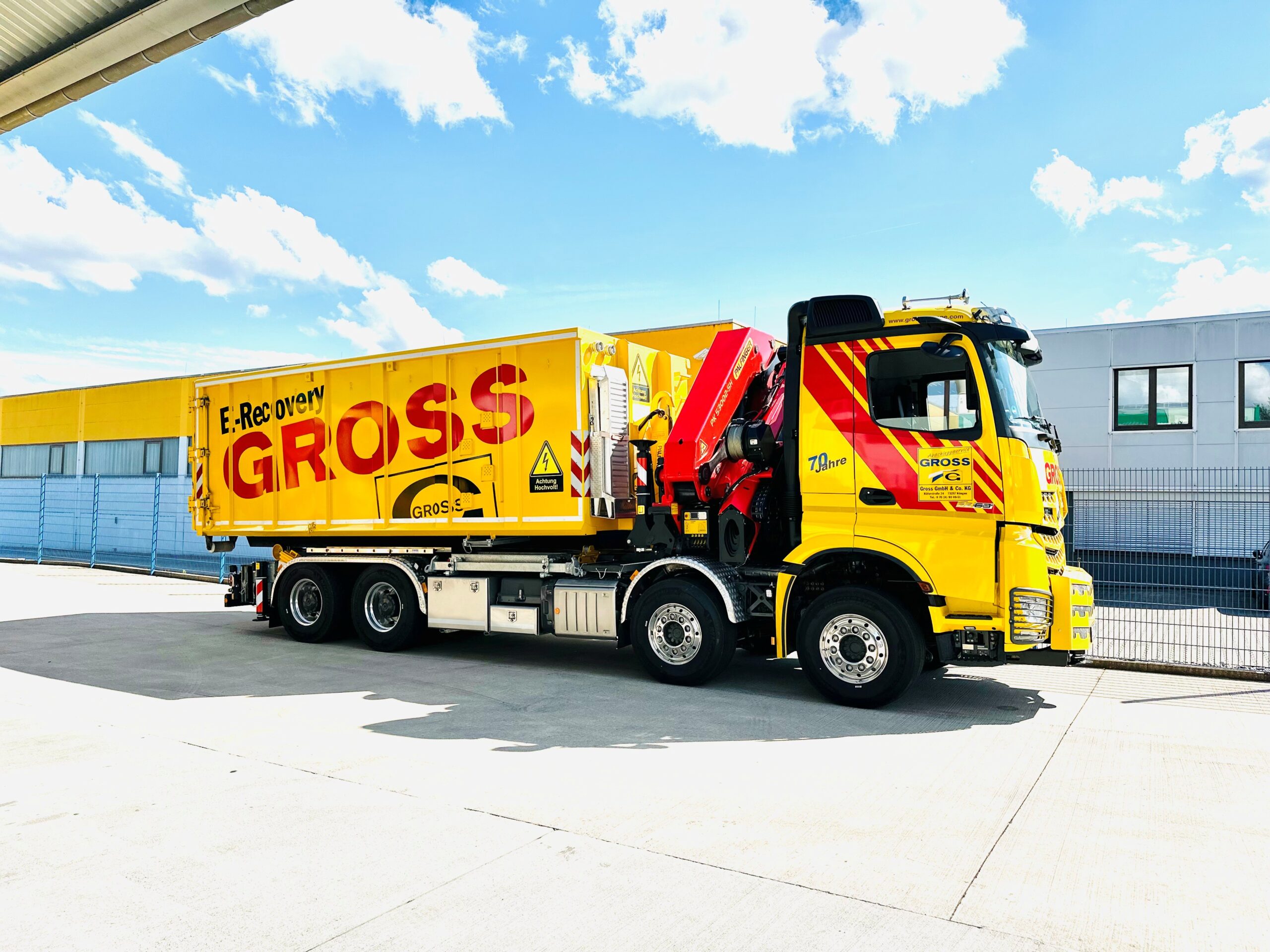 Gelber Lastwagen mit „E-Recovery Gross“ auf dem Container, steht auf einem sonnigen Industriegelände neben einem Gebäude.