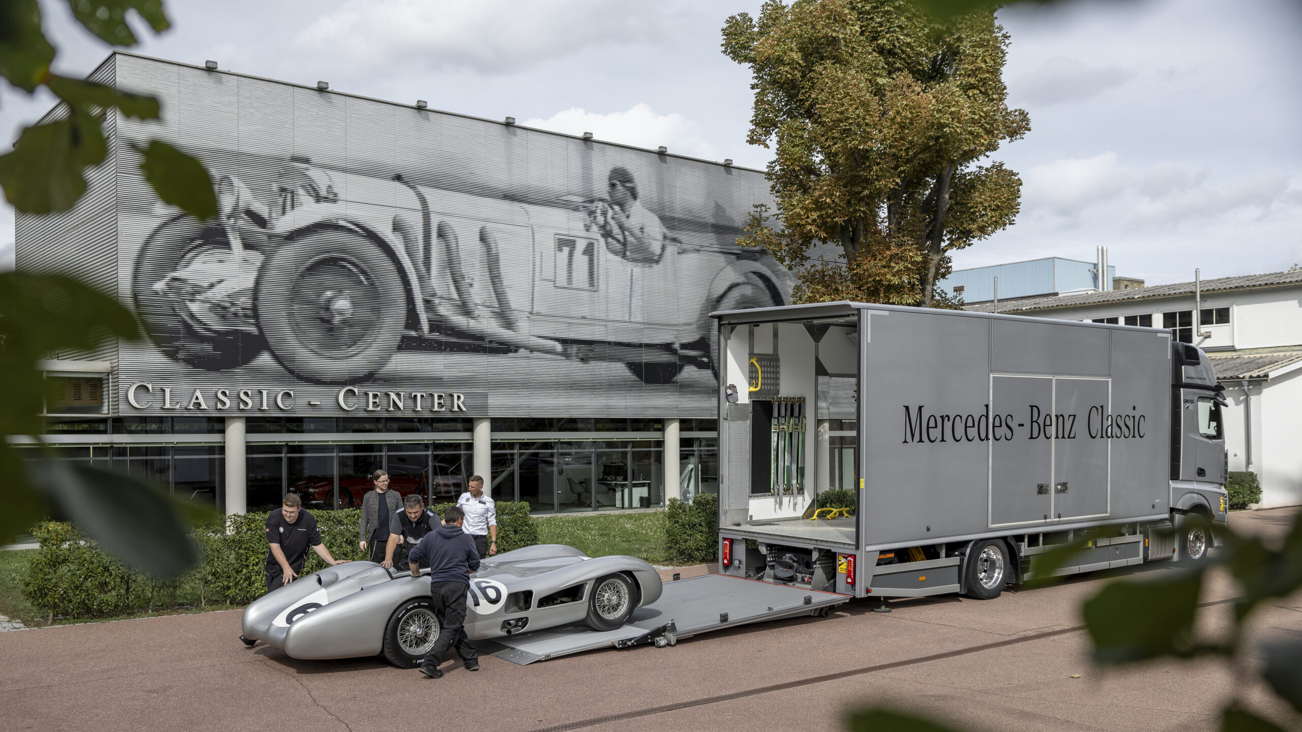 Historisches Mercedes-Rennauto wird vor einem LKW am Mercedes-Benz Classic Center ausgeladen. Große Wandkunst eines Rennwagens.