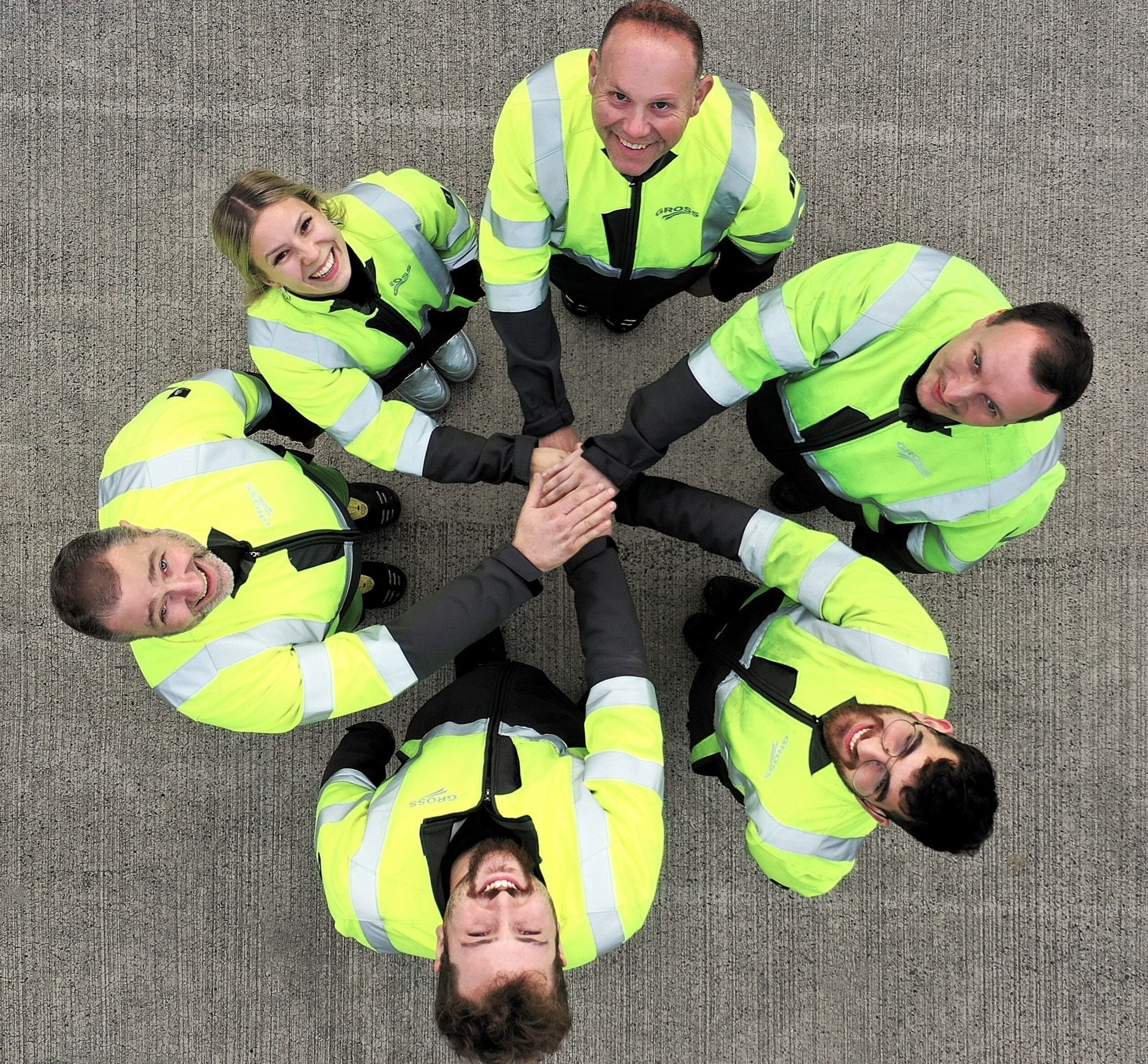Sechs Personen in gelben Warnwesten bilden einen Kreis und legen die Hände zusammen, lächelnd auf grauem Boden.