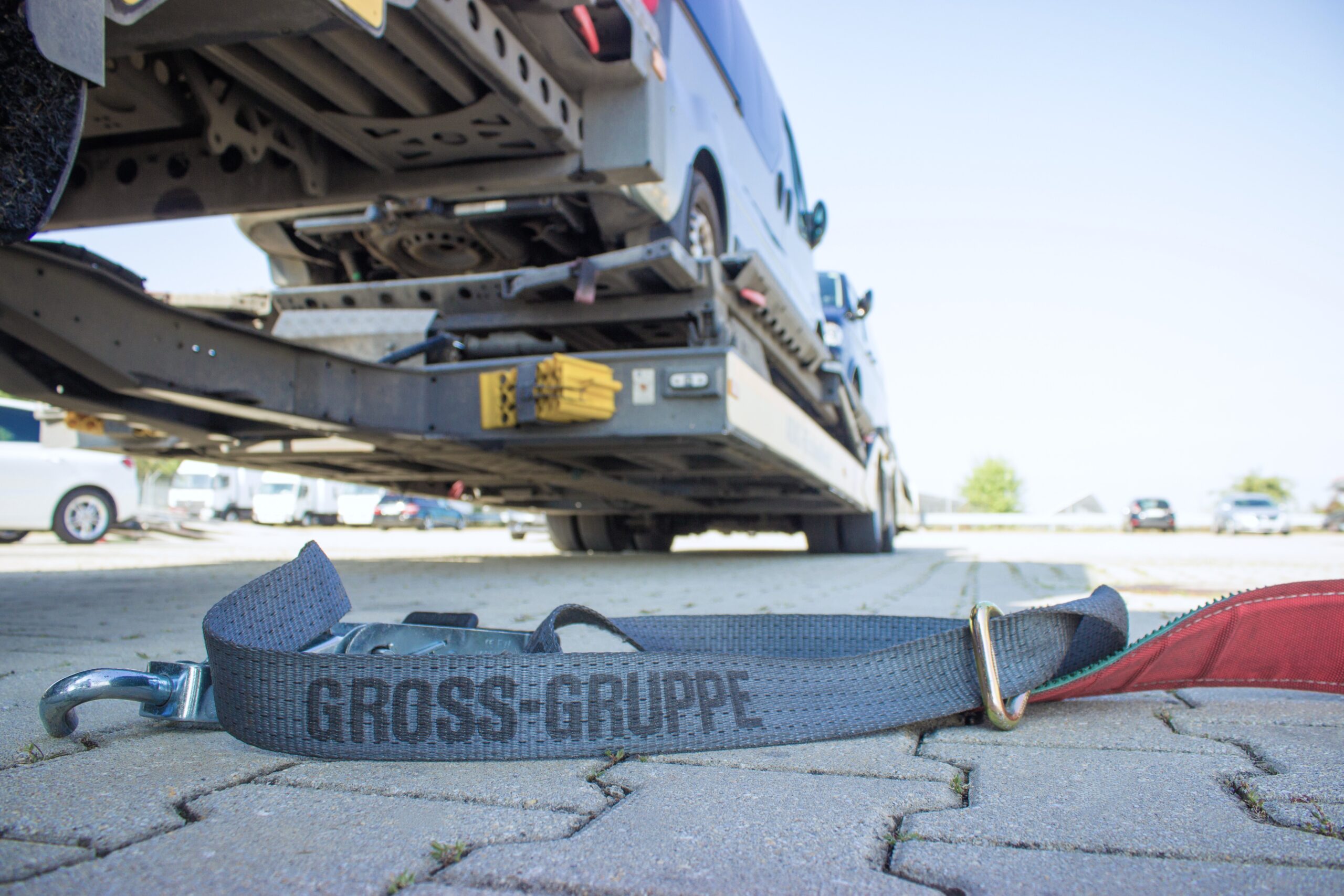Transportanhänger mit Auto darauf, im Vordergrund ein Spanngurt mit Aufschrift Grossi-Gruppe auf gepflastertem Boden.