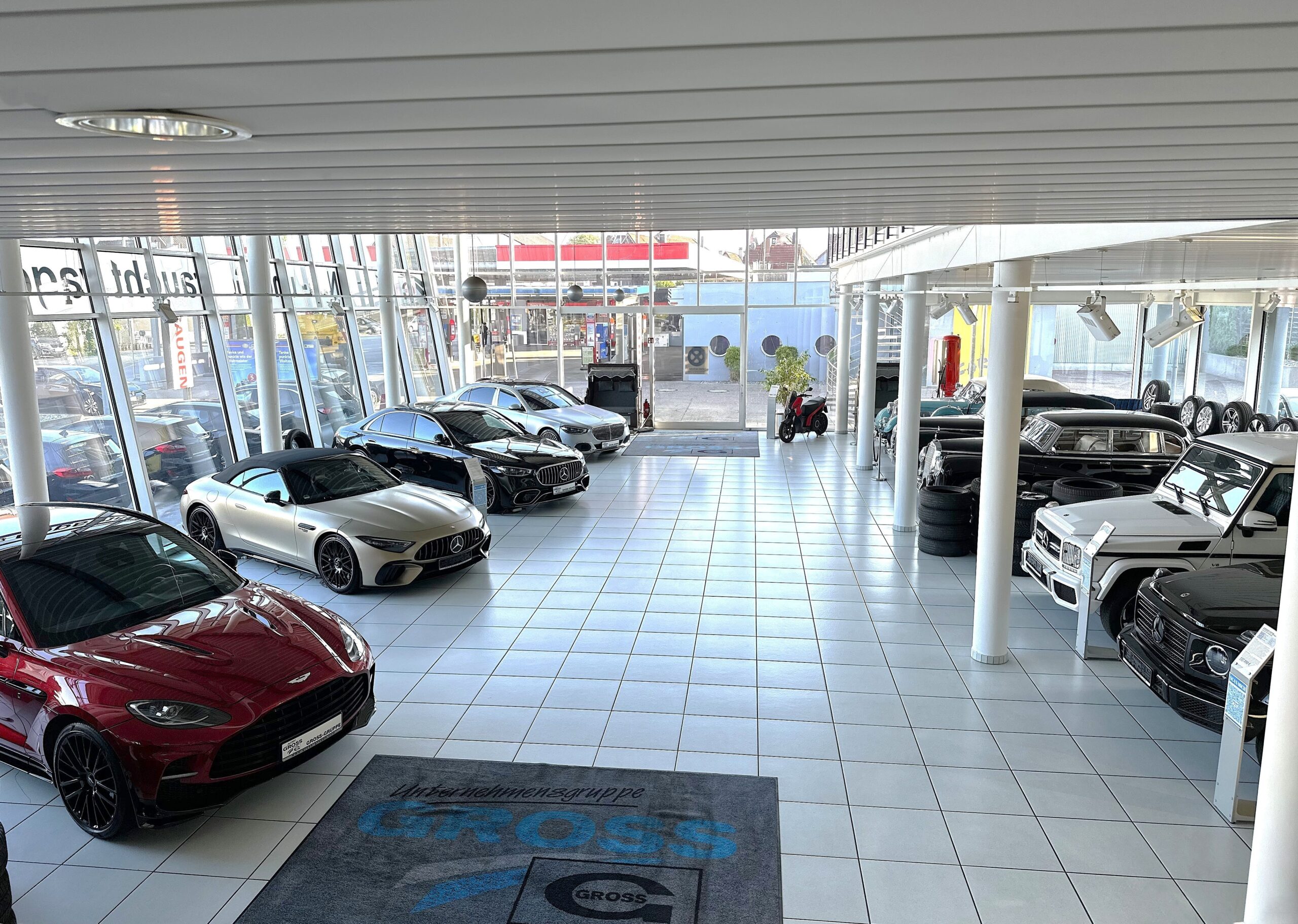 Autohaus-Showroom mit verschiedenen Luxusfahrzeugen auf glänzenden Fliesen, von großen Fenstern umgeben.