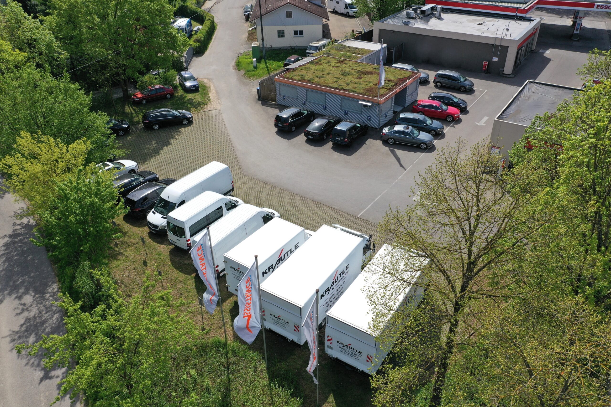 Luftaufnahme eines Parkplatzes mit weißem Lieferwagen, parkenden Autos und begrüntem Dach eines Gebäudes, umgeben von Bäumen.