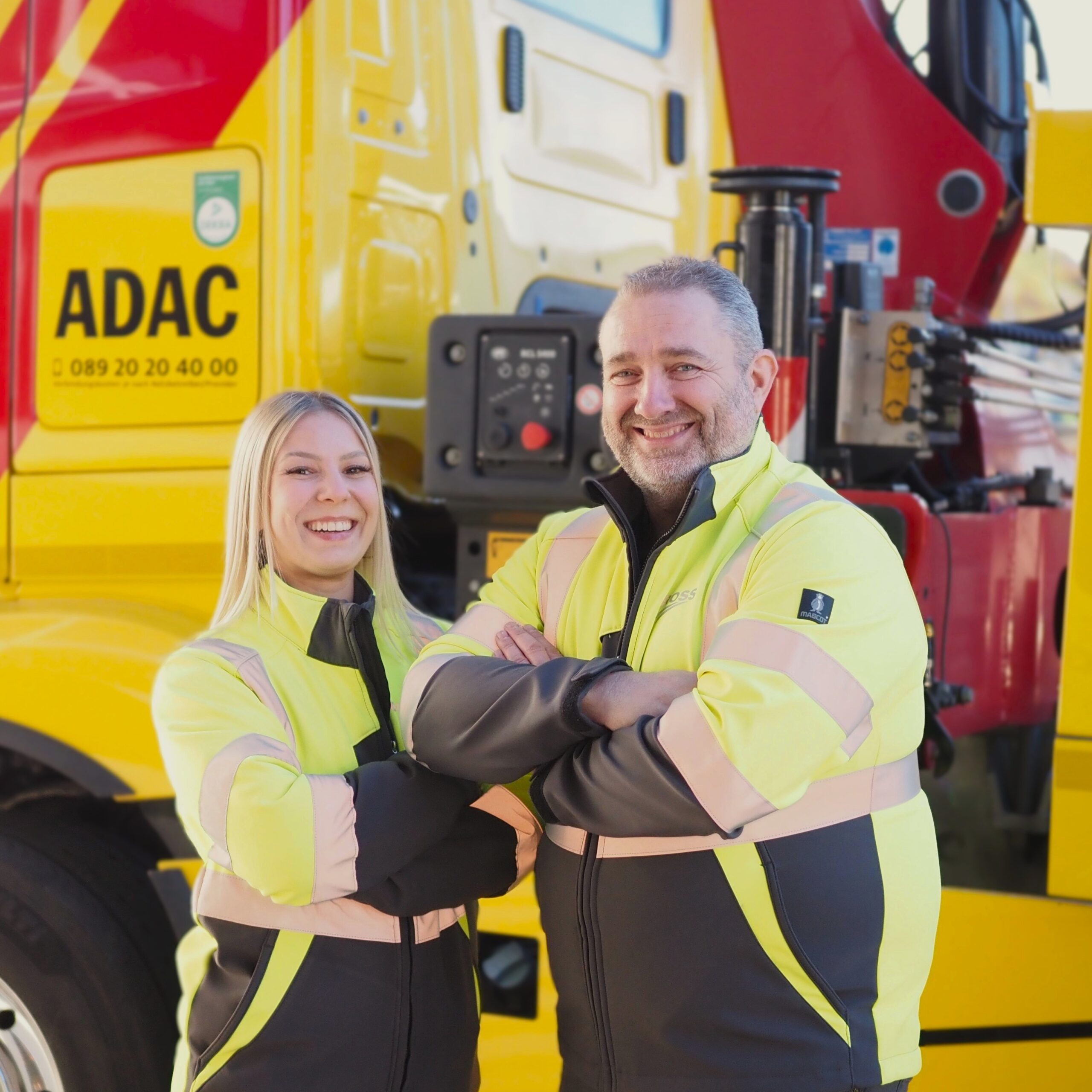 Zwei lächelnde Personen in gelber Arbeitskleidung stehen vor einem gelben ADAC-Fahrzeug.
