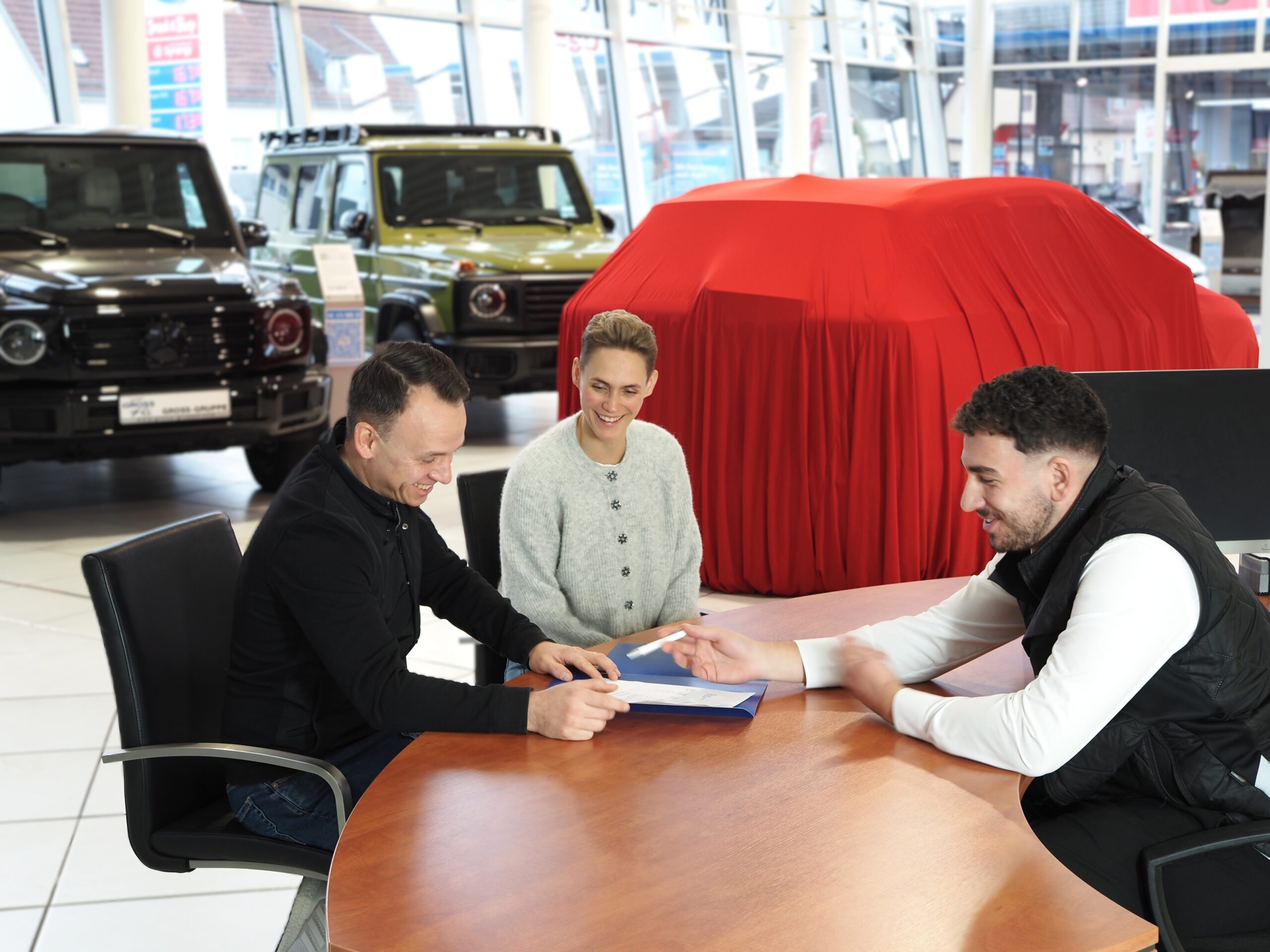 Drei Personen im Autohaus, unterzeichnen Dokumente am Tisch, Autos im Hintergrund, eines rot verhüllt.