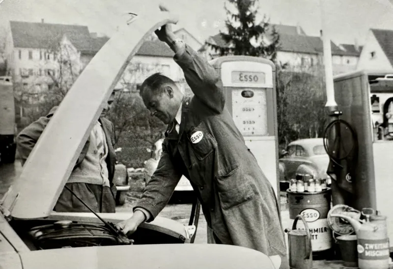 Mechaniker öffnet Motorhaube eines Oldtimers an einer Esso-Tankstelle in historischer Schwarz-Weiß-Aufnahme.
