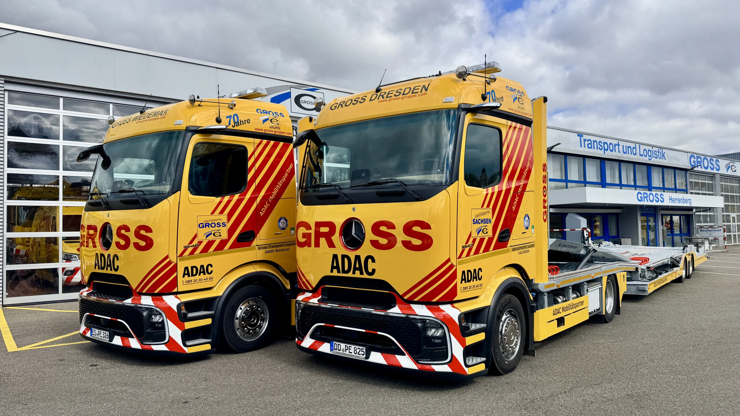 Zwei gelbe Lkw mit ADAC-Logos stehen vor einem modernen Logistikgebäude, umgeben von Wolken.