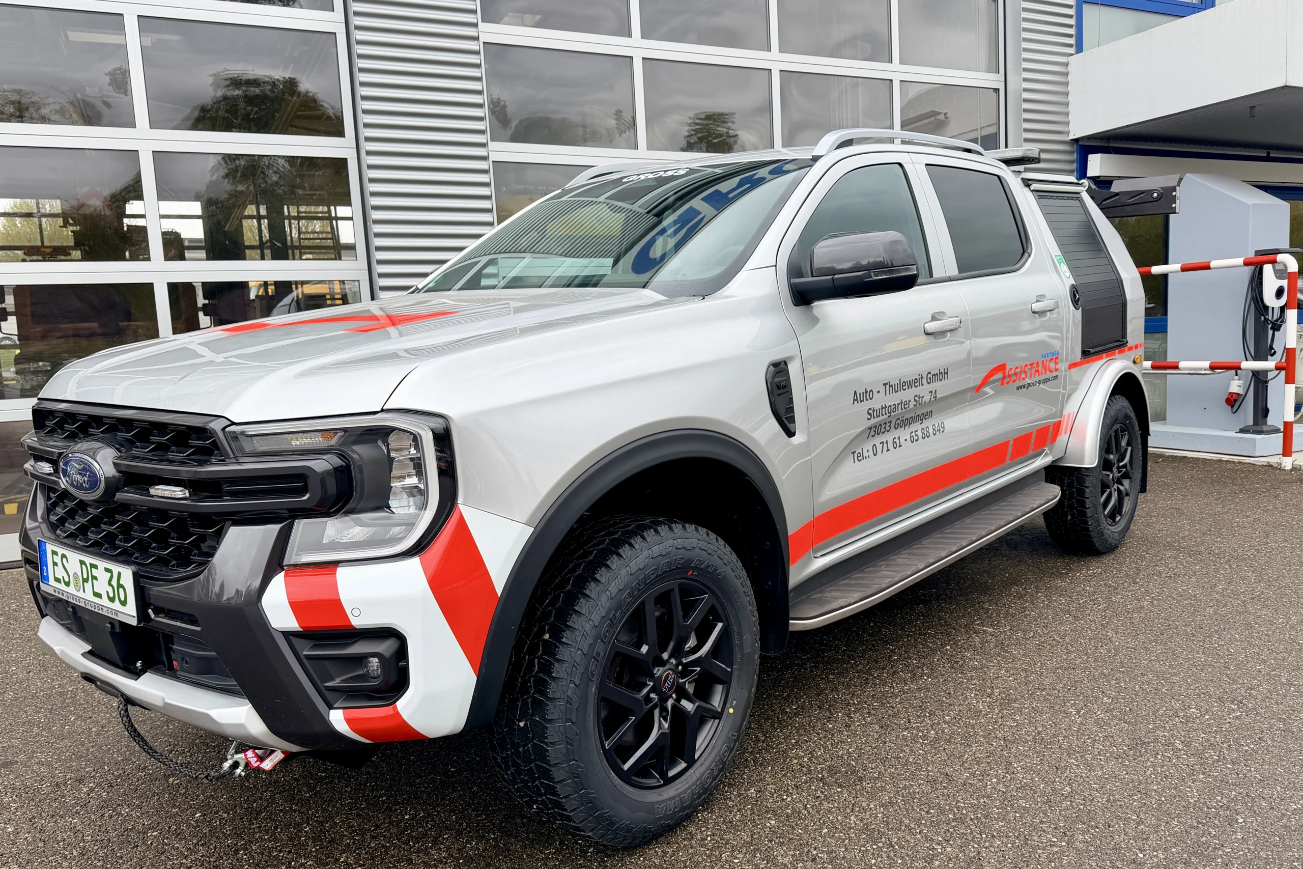 Grauer SUV mit roten und weißen Streifen, geparkt vor einem Gebäude. Schriftzüge auf der Seite und offroad-tauglichen Reifen.